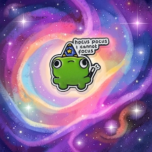 Puede incluir: Pin de rana verde con sombrero de mago y varita mágica, sobre un fondo de galaxia colorida. El pin tiene el texto "hocus pocus I cannot focus" en un bocadillo.