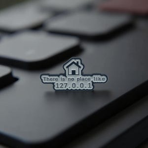 Könnte beinhalten: Ein silberner und schwarzer Emaille-Pin mit dem Text "There is no place like 127.0.0.1" und einer Hausgrafik. Der Pin befindet sich auf einer dunkelgrauen Oberfläche, möglicherweise einer Tastatur.