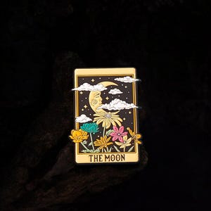 Può includere: Spilla rettangolare con un design di carta dei tarocchi e un bordo dorato. La carta raffigura una luna crescente con un volto, nuvole, fiori e le parole "THE MOON". I fiori sono gialli, turchese e rosa.