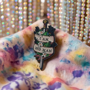 Könnte beinhalten: Dekorative Anstecknadel mit einem Schwertdesign und dem Text "I AM NO MAN" auf einem Banner. Die Nadel ist mit weißen Blumen, grünen Blättern und schwarzen Akzenten verziert, vor einem farbenfrohen Batik-Hintergrund.