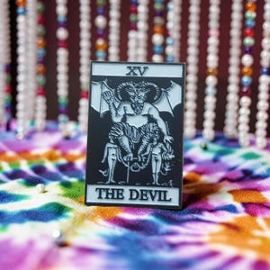 Könnte beinhalten: Ein schwarz-weißer Emaille-Pin mit der Tarotkarte Der Teufel. Der Pin zeigt eine gehörnte Figur mit Flügeln, die sitzt und zwei Figuren hält. Der Text "XV" und "THE DEVIL" sind auf dem Pin abgebildet.