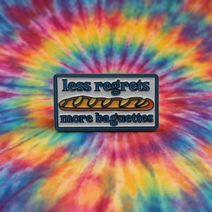 Könnte beinhalten: Rechteckiger Anstecker mit blauem Rand und den Worten "less regrets" über einer Baguette-Illustration und den Worten "more baguettes". Der Anstecker befindet sich vor einem bunten Batik-Hintergrund.
