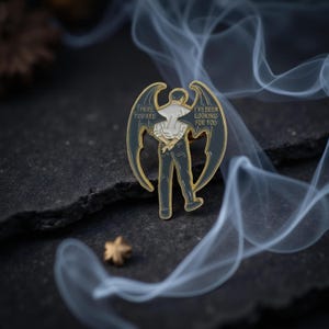 Könnte beinhalten: Ein schwarz-goldener Emaille-Pin mit einer Figur mit dunklen Flügeln. Die Figur trägt ein weißes Hemd und eine dunkle Hose. Auf den Flügeln steht der Text "THERE YOU ARE" und "I'VE BEEN LOOKING FOR YOU".