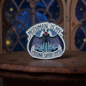 Könnte beinhalten: Runder Emaille-Pin mit einem Mothman-Design. Der Pin zeigt den Text "Mothman is my Emotional Support Cryptid". Der Mothman hat rote Augen und ist vor einem blau-violetten Hintergrund platziert. Der Pin ist schwarz und weiß.