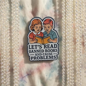 May include: A colorful pin featuring two children reading a book, with the text "LET'S READ BANNED BOOKS - AND CAUSE PROBLEMS." The pin has a vintage, retro design with a white border and is set against a patterned background.