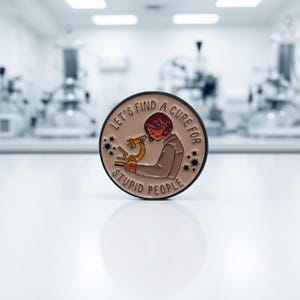 Könnte beinhalten: Runder Emaille-Pin mit dem Text "LET'S FIND A CURE FOR STUPID PEOPLE". Der Pin zeigt eine Cartoon-Person, die ein Mikroskop benutzt. Der Pin hat einen hellbraunen Hintergrund mit schwarzen und goldenen Akzenten.