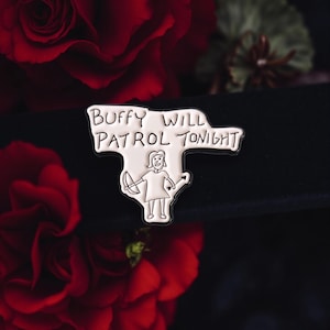 Könnte beinhalten: Ein silberfarbener Emaille-Pin mit dem Text "Buffy Will Patrol Tonight" und einer Illustration einer Figur mit Pfeil und Bogen. Der Pin befindet sich vor einem dunklen Hintergrund mit roten Rosen.