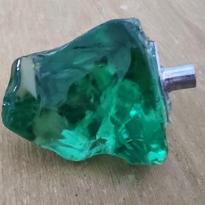 May include: A decorative cabinet knob with a unique, organic shape. The knob is made of translucent, emerald green resin with a silver-colored metal stem. The knob's irregular form gives it a natural, handcrafted appearance.