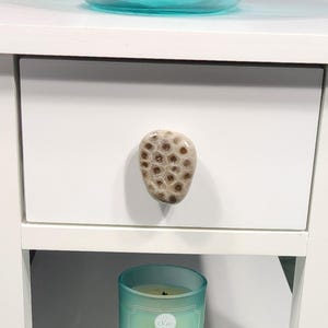 May include: A white nightstand with a coral-shaped drawer pull. A teal glass candle with the text "Calming Waves" sits below the drawer. A teal glass bowl is on top of the nightstand.