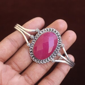 Puede incluir: Un brazalete de plata con una gran piedra preciosa magenta facetada de forma ovalada en el centro. El engaste de metal muestra intrincados detalles de cuentas y alambre trenzado alrededor de la piedra, con un dise&ntilde;o de banda de v&aacute;stago dividido.