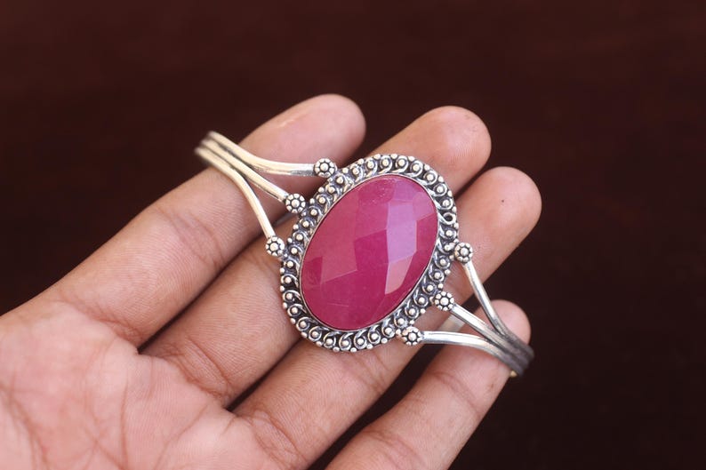Puede incluir: Un brazalete de plata con una gran piedra preciosa magenta facetada de forma ovalada en el centro. El engaste de metal muestra intrincados detalles de cuentas y alambre trenzado alrededor de la piedra, con un dise&ntilde;o de banda de v&aacute;stago dividido.