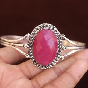 May include: A silver cuff bracelet featuring a large, oval-shaped, faceted pink gemstone at the center. The metal setting displays intricate beaded and twisted wire detailing surrounding the stone, with a split band design extending to the sides.