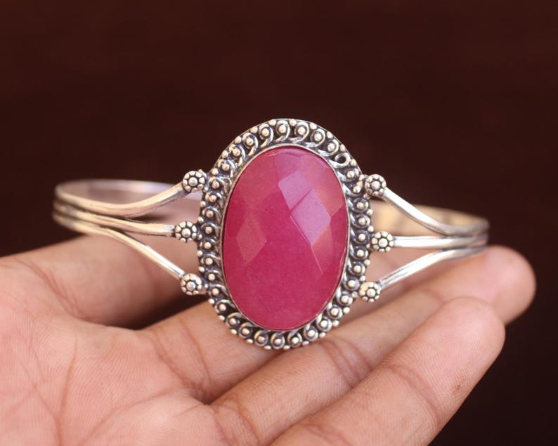 Puede incluir: Un brazalete de plata con una gran piedra preciosa rosa facetada de forma ovalada en el centro. El engaste de metal muestra intrincados detalles de cuentas y alambre trenzado alrededor de la piedra, con un dise&ntilde;o de banda dividida hacia los lados.