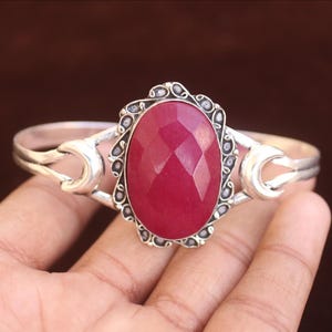 Op de afbeelding: Een zilveren armband met een grote, ovale, gefacetteerde donkerroze edelsteen in het midden. De steen is gezet in een decoratieve, geschulpte zilveren rand en de band heeft aan beide zijden accenten in de vorm van een halve maan.