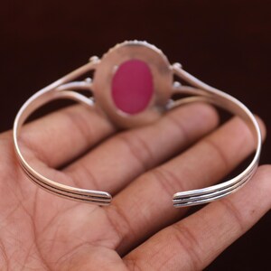 Puede incluir: Una pulsera tipo brazalete de tono plateado con una piedra rosa de forma ovalada en el centro. La banda met&aacute;lica tiene un dise&ntilde;o dividido que converge cerca del engaste de la piedra, con tres l&iacute;neas horizontales paralelas en los lados.