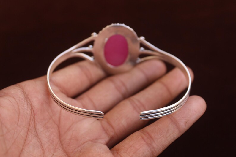 Puede incluir: Una pulsera tipo brazalete de tono plateado con una piedra rosa de forma ovalada en el centro. La banda met&aacute;lica tiene un dise&ntilde;o dividido que converge cerca del engaste de la piedra, con tres l&iacute;neas horizontales paralelas en los lados.