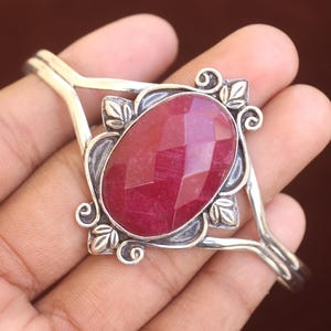 Puede incluir: Un brazalete de plata con una gran piedra preciosa de color rojo oscuro, forma ovalada y tallada en facetas en el centro. El engaste de metal muestra intrincados motivos de volutas y hojas alrededor de la piedra, con un diseño de banda dividida.