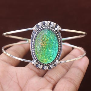 May include: A silver cuff bracelet featuring a large, oval-shaped iridescent green stone at the centre. The stone is surrounded by a decorative metal border with small flower-like motifs and beaded detailing. The band consists of two thin silver wires.