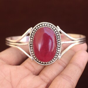 Può includere: Un bracciale a fascia in argento con una grande pietra preziosa ovale di colore rosso scuro e superficie liscia al centro. La pietra è incorniciata da un bordo decorativo in argento perlato e la fascia si divide in due bracci sottili in argento.