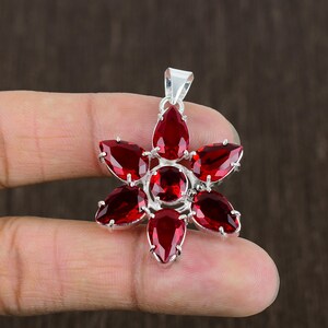Puede incluir: Un colgante de plata con un diseño de flor de piedra preciosa roja. El colgante presenta una piedra preciosa redonda central rodeada de seis piedras preciosas en forma de pera.