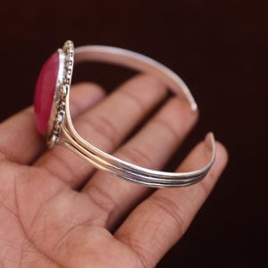 Puede incluir: Un brazalete de plata con una piedra grande, ovalada, lisa y de color rosa intenso, engastada en un marco de plata decorativo con cuentas. La banda del brazalete est&aacute; formada por tres hilos de plata paralelos que se unen en los extremos.