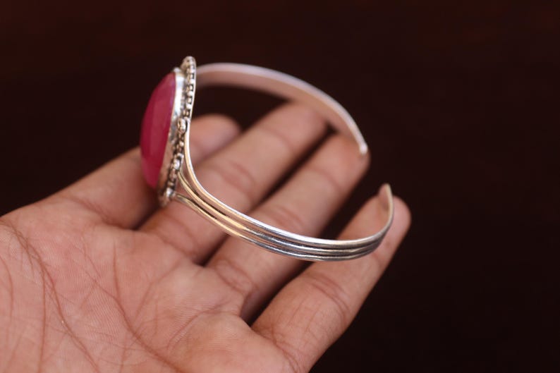 Puede incluir: Un brazalete de plata con una piedra grande, ovalada, lisa y de color rosa intenso, engastada en un marco de plata decorativo con cuentas. La banda del brazalete est&aacute; formada por tres hilos de plata paralelos que se unen en los extremos.