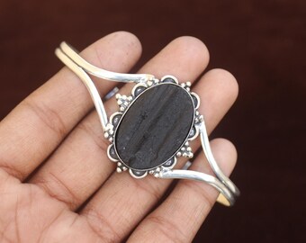Puede incluir: Un brazalete de plata con una piedra negra grande, ovalada y texturizada en el centro. La piedra está enmarcada por un engaste de plata decorativo con pequeños detalles en forma de cuentas. La banda del brazalete se divide en dos brazos de plata pulida.