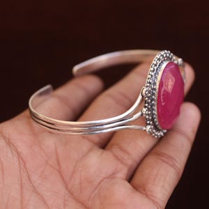 Puede incluir: Un brazalete de plata con una gran piedra preciosa de color rosa intenso, de forma ovalada y facetada, engastada en un marco de plata decorativo con cuentas. La banda del brazalete consta de tres hilos de plata finos y paralelos que se unen.
