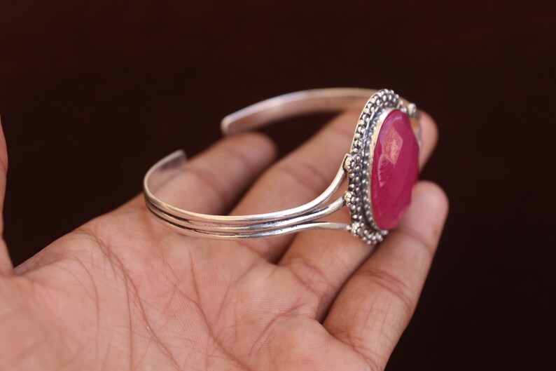 Puede incluir: Un brazalete de plata con una gran piedra preciosa de color rosa intenso, de forma ovalada y facetada, engastada en un marco de plata decorativo con cuentas. La banda del brazalete consta de tres hilos de plata finos y paralelos que se unen.