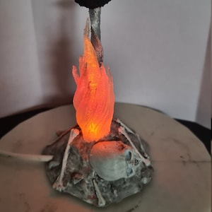 May include: A decorative lamp featuring a sword with a brown handle and a ring-shaped pommel. The sword is set above a base with a skull, bones, and a glowing orange flame. The lamp is on a white circular base.
