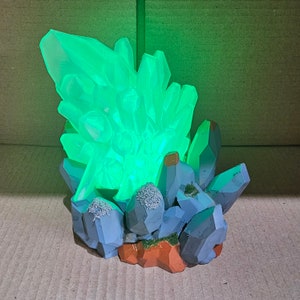May include: A glowing green crystal formation sculpture. The crystal cluster is atop a base of blue and gray rock formations with patches of green moss and orange-brown earth. The crystal emits a bright, vibrant green light.