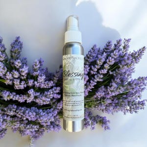 May include: A silver spray bottle labeled "Blessing Smudge Spray" with a white nozzle, set against a backdrop of purple flowers. The bottle's label includes the text "Sage Lady Trading Co."