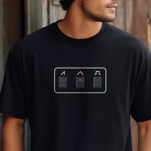 May include: A black t-shirt with a white graphic of three black buttons with red lights. The buttons are labeled with the letters "I", "^", and "L".