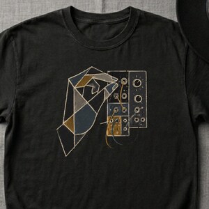 May include: Black t-shirt featuring a geometric design of a hand interacting with a control panel. The graphic uses shades of brown, blue, and gold. The shirt is paired with blue jeans, a black hat, and sunglasses.