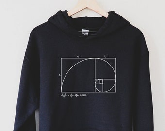 Puede incluir: Sudadera con capucha negra con un gráfico blanco de la proporción áurea, un concepto matemático representado por una espiral dentro de un cuadrado. La ecuación de la proporción áurea está impresa debajo del gráfico.