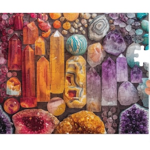May include: A colourful jigsaw puzzle depicting various gemstones in shades of orange, yellow, purple, and blue. The puzzle pieces are arranged to resemble the gemstones. A hand holds a puzzle piece, ready to complete the image.