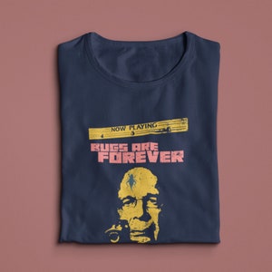 May include: A navy blue t-shirt with a graphic of a man's face with a bug on his head. The text "NOW PLAYING" and "BUGS ARE FOREVER" is printed on the shirt.