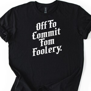 Op de afbeelding: Zwart T-shirt met de witte tekst "Off To Commit Tom Foolery". Het shirt is opgevouwen, met opgerolde mouwen. De achtergrond is effen wit.