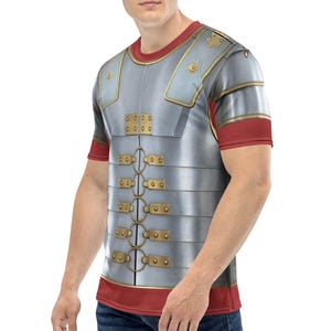 May include: A short-sleeved t-shirt featuring a detailed print of Roman armor. The design includes a silver-colored breastplate with gold-colored accents and red trim on the sleeves and neckline. The shirt is worn by a person.