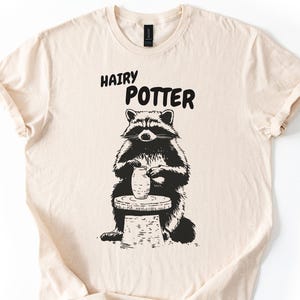 May include: A cream-coloured t-shirt featuring the text "HAIRY POTTER" above a black and white illustration of a raccoon at a pottery wheel, holding a vase. The raccoon is wearing a black and white outfit.