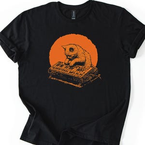 May include: Black t-shirt featuring an orange illustration of a cat playing a keyboard against an orange circle background. The cat is depicted in a detailed, vintage style. The shirt is made of a soft material.
