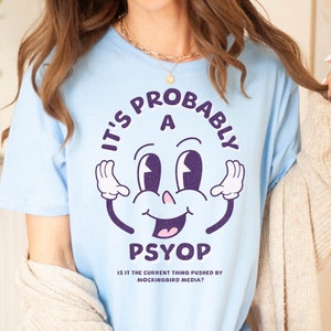 May include: A light blue t-shirt with a cartoon face and the text "It's probably a psyop. Is it the current thing pushed by mockingbird media?"