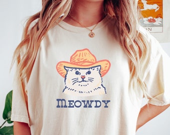 May include: A white t-shirt with a cartoon image of a cat wearing a cowboy hat. The text "MEOWDY" is printed below the cat.