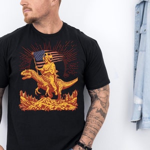 Puede incluir: Camiseta negra con un gráfico de una persona montando un dinosaurio con una bandera estadounidense. El gráfico está en tonos de naranja, amarillo y rojo.