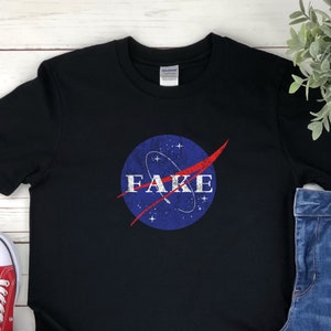 K&ouml;nnte beinhalten: Ein schwarzes T-Shirt mit einem abgenutzten blau-roten NASA-Logo, auf dem in wei&szlig;er Schrift "FAKE" steht.