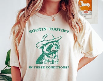 Puede incluir: Una camiseta de color crema con un mapache verde que lleva un sombrero de vaquero y el texto "Rootin' Tootin'? In these conditions?"
