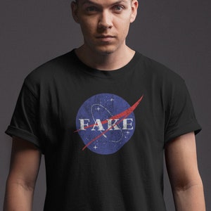 May include: A black t-shirt with a blue and red NASA-inspired logo that says "FAKE" in white letters.