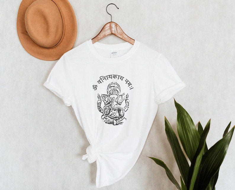 May include: White t-shirt with a black and white illustration of Ganesha, a Hindu deity, with the text "ॐ वनितायकाय नमः!" in Devanagari script.