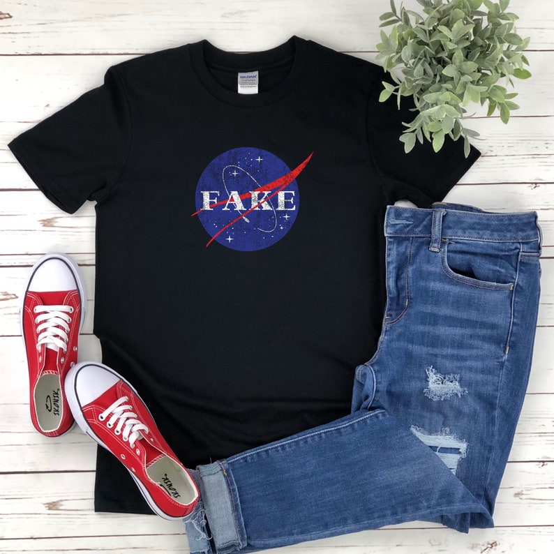 K&ouml;nnte beinhalten: Ein schwarzes T-Shirt mit einem abgenutzten blau-roten NASA-Logo, auf dem in wei&szlig;er Schrift "FAKE" steht.