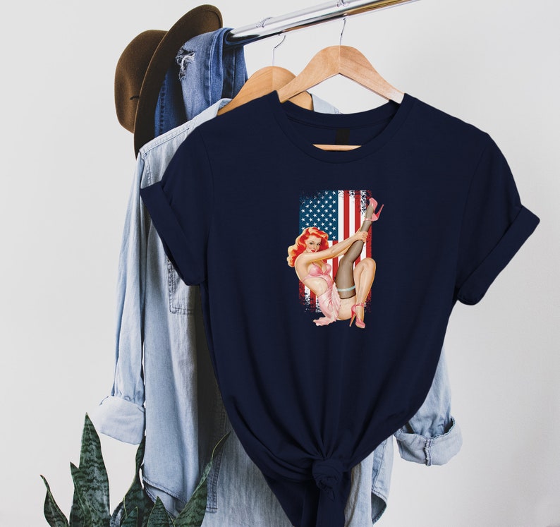 May include: A navy blue t-shirt with a vintage pin-up girl illustration wearing red heels and a pink dress, posed in front of an American flag.